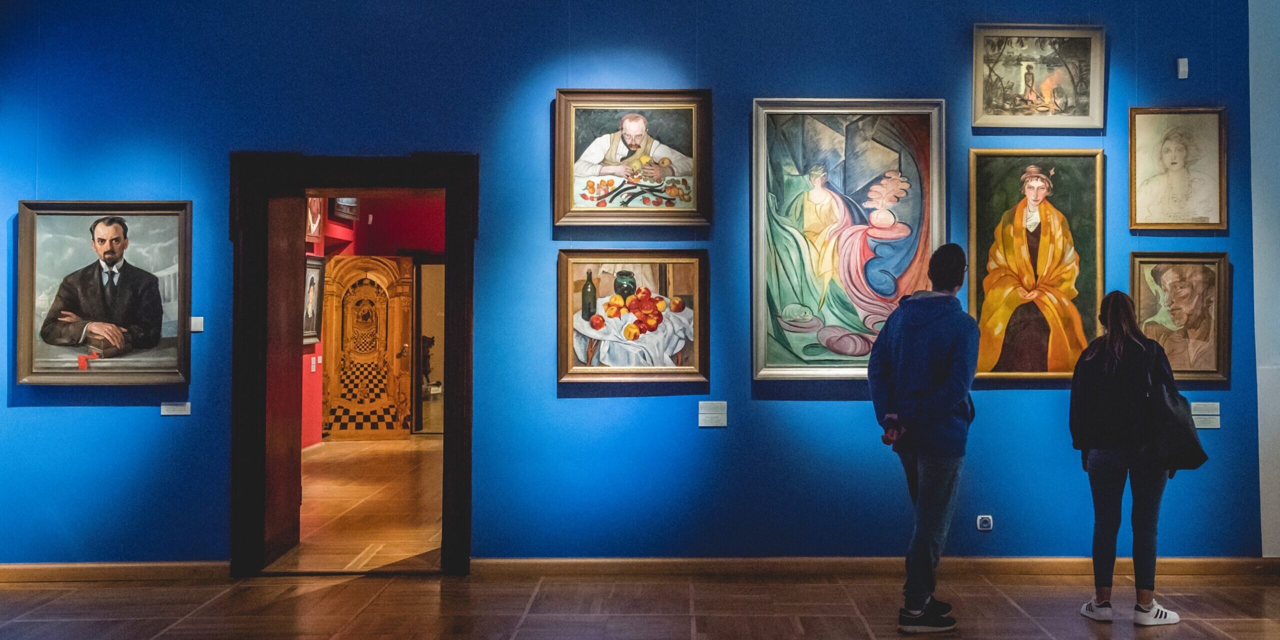 Fotografia przedstawia dwoje ludzi ujetych od tyłu oglądających obrazy zawieszone na ścianie galerii.