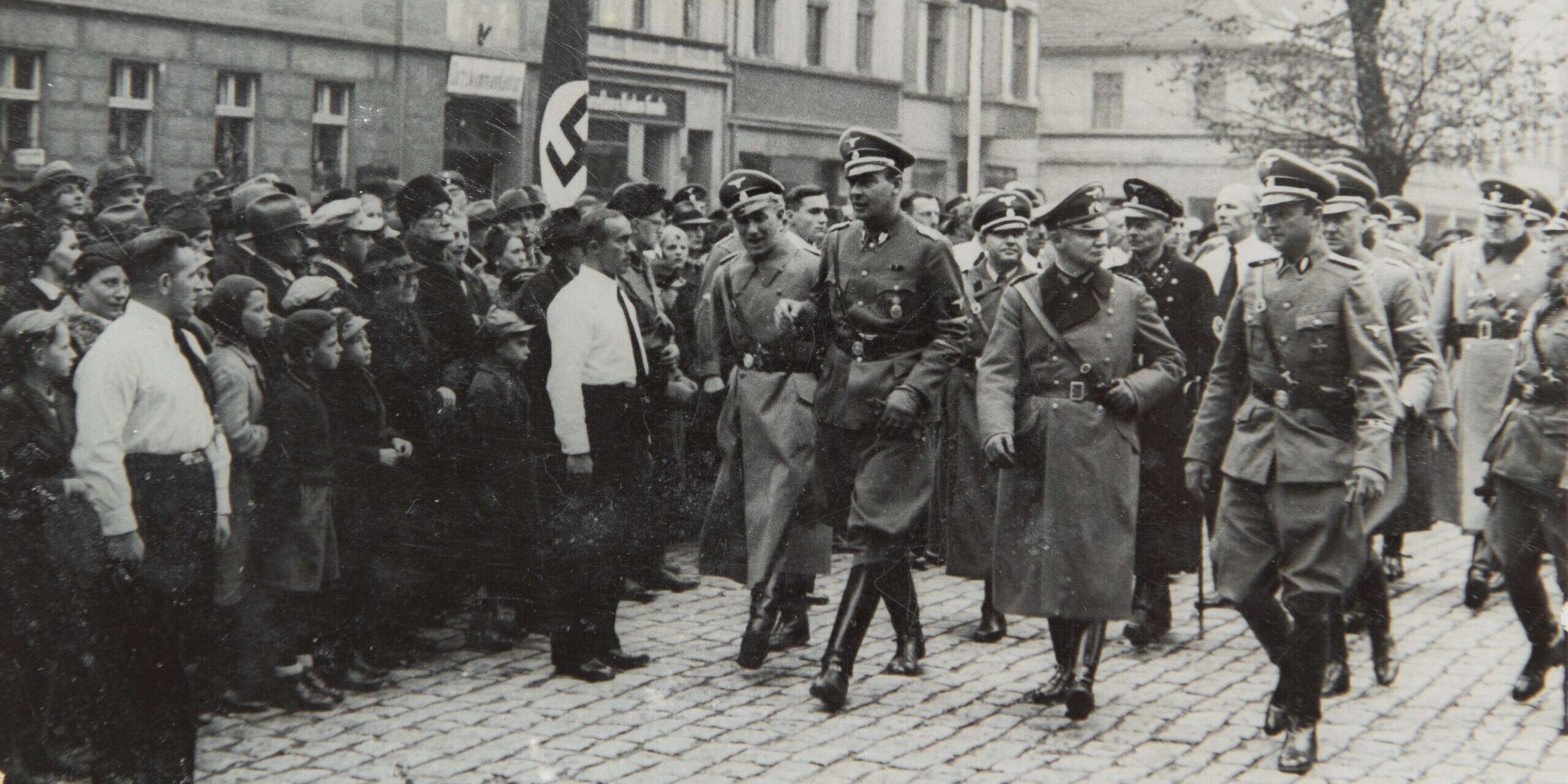 Fotografia przedstawia grupę oficerów niemieckich z II wojny światowej maszerujących przed tłumem ludzi.