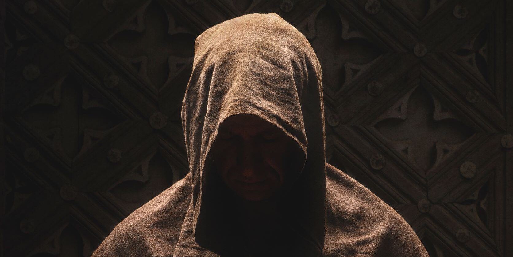 Fotografia przedstawia postać zakonnika w kapturze na ciemnym tle.