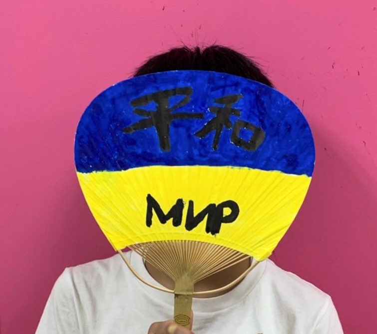 Zdjęcie przedstawia dziecko - ucznia japońskiej szkoły kaligraficznej. Dziecko zakryło swoją twarz wachlarzem "uchiwa" (wachlarz sztywny, okrągły, nieskładany) z kaligrafiami, który został pomalowany w niebiesko-żółte barwy, barwy flagi ukraińskiej.