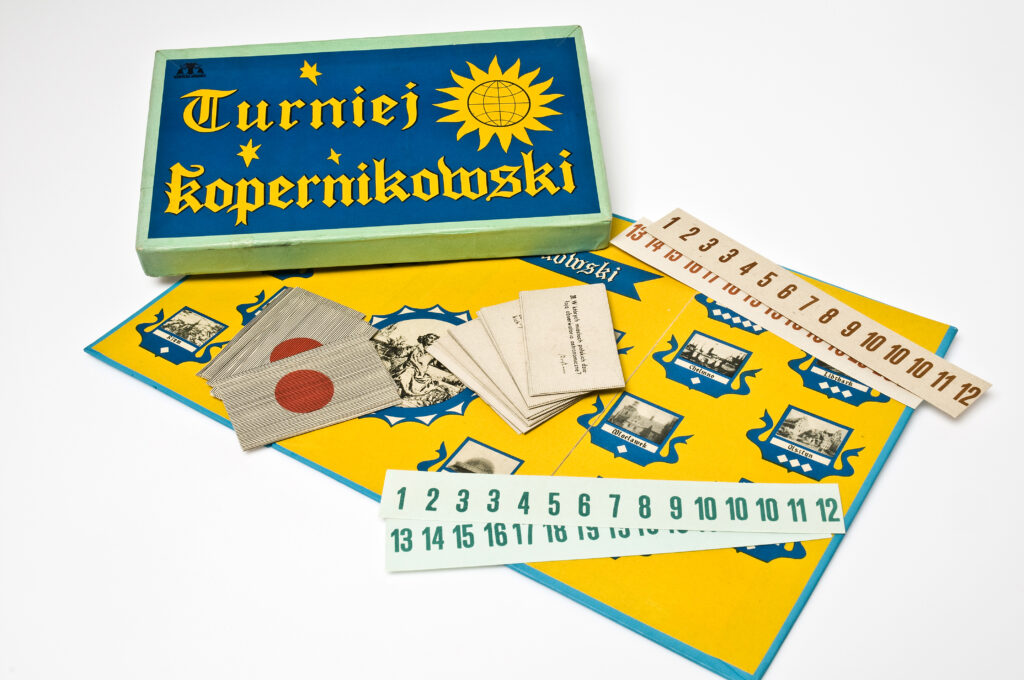 Zdjęcie przedstawia grę planszową "Turniej kopernikowski". Na żółtej planszy gry, leżą jej elementy, m.in kart do gry.