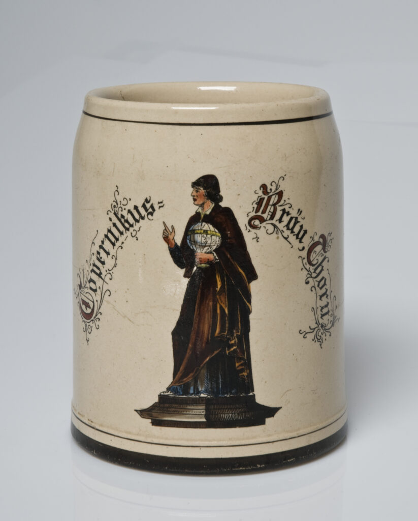Zdjęcie przedstawia kubek najprawdopodobniej gliniany z wizerunkiem pomnika Mikołaja Kopernika oraz napisem z języku niemieckim. 