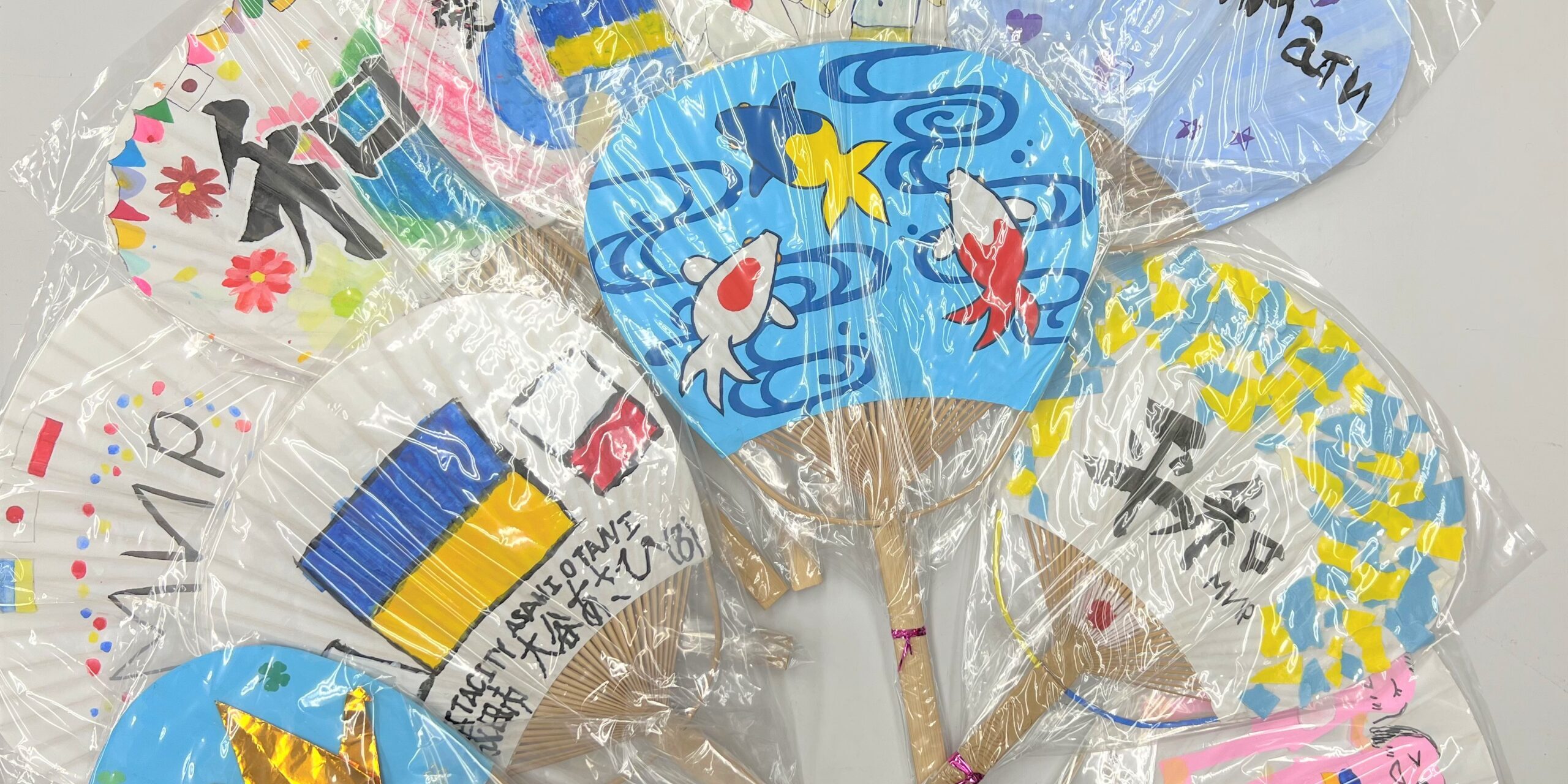 Obrazek przedstawia kilka ułożonych obok siebie wachlarzy "uchiwa". Wachlarze są bardzo kolorowe, namalowane są na nich symbole barw narodowych Japonii i Polski (biało czerwone) oraz Ukrainy (niebiesko-żółte).