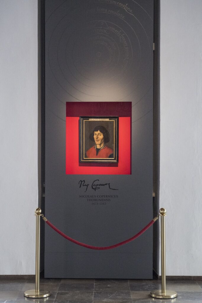 Fotografia portretu Mikołaja Kopernika, znajdującego się w Sali Mieszczańskiej Ratusza Staromiejskiego w Toruniu. Prezentowana w nowej odsłonie. Chroniona szybą, w specjalnej, pionowej ściance. Wnętrze czerwone, na zewnątrz szara, a na górze zarysowany układ heliocentryczny zaczerpnięty z dzieła Mikołaja Kopernika.