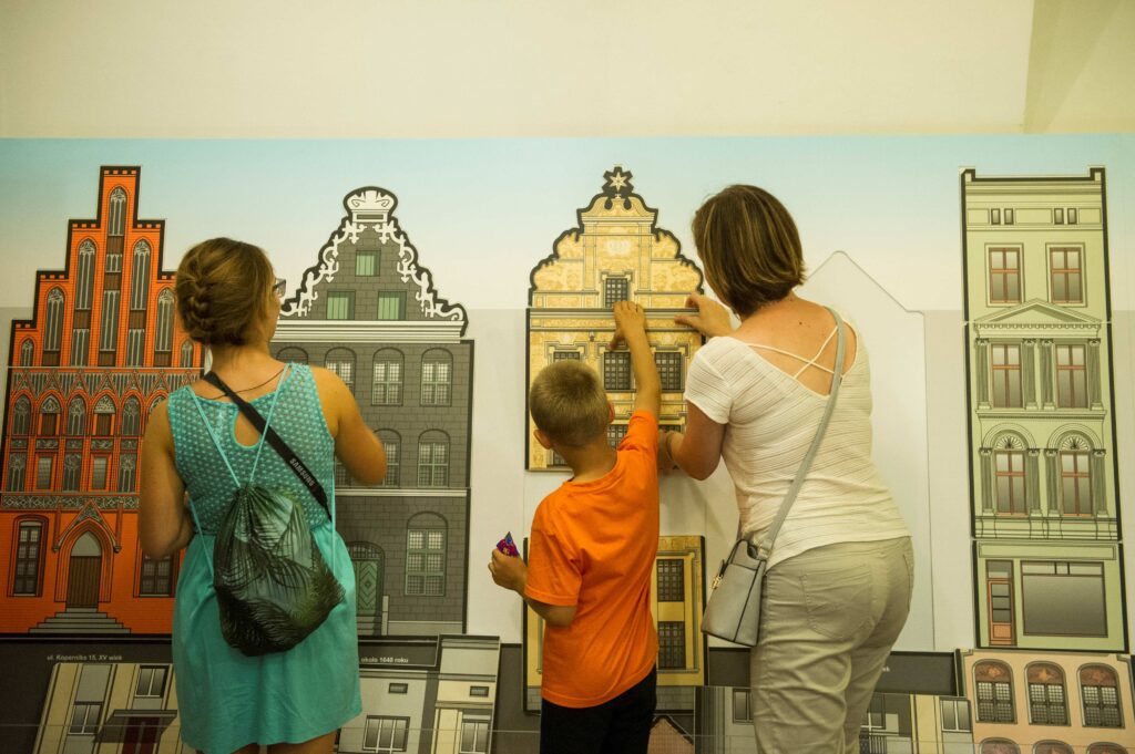 Dwie osoby dorosłe i dziecko, widziane od tyłu, układają puzzle wielkogabarytowe