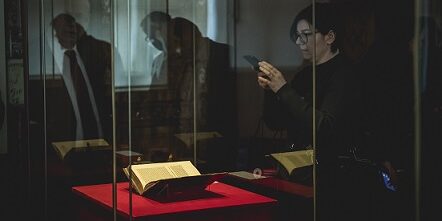 Szklana gablota, w której zabezpieczona jest książka „De revolutionibus orbium coelestium” wyd. 1 Mikołaja Kopernika. Zdjęcie w tajemniczej, ciemnje tonacji. Po prawej stronie zwiedzająca, która fotografuje księgę telefonem.