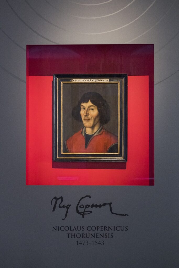 Portret Mikołaja Kopernika (tzw. gimnazjalny). Obraz umieszczony za szybą, zawieszony na czerwonym tle. 