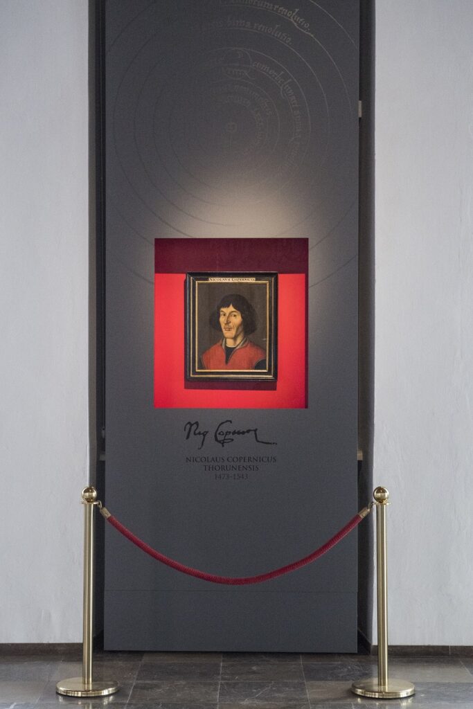 Portret Mikołaja Kopernika (tzw. gimnazjalny). Obraz umieszczony za szybą, zawieszony na czerwonym tle. 