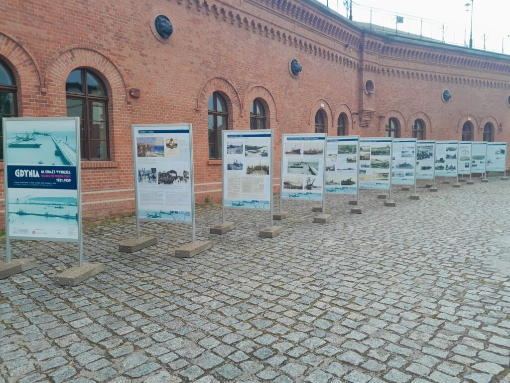 Wystawa planszowa ustawiona na dziedzińcu przed Muzeum Twierdzy Toruń - każda plansza stoi osobno. Z tyłu frontalna, ceglana ściana muzeum. 