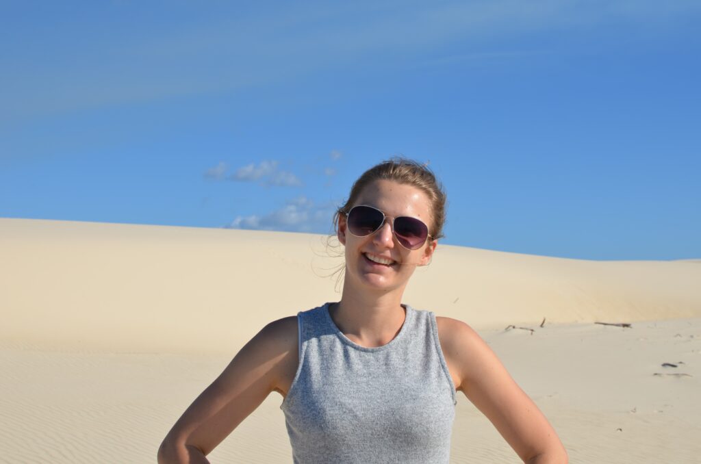 W centralnej części uśmiechnięta młoda kobieta w okularach przeciwsłonecznych widziana od piersi w górę, w tle piaszczysta plaża, u góry niebieskie niebo.