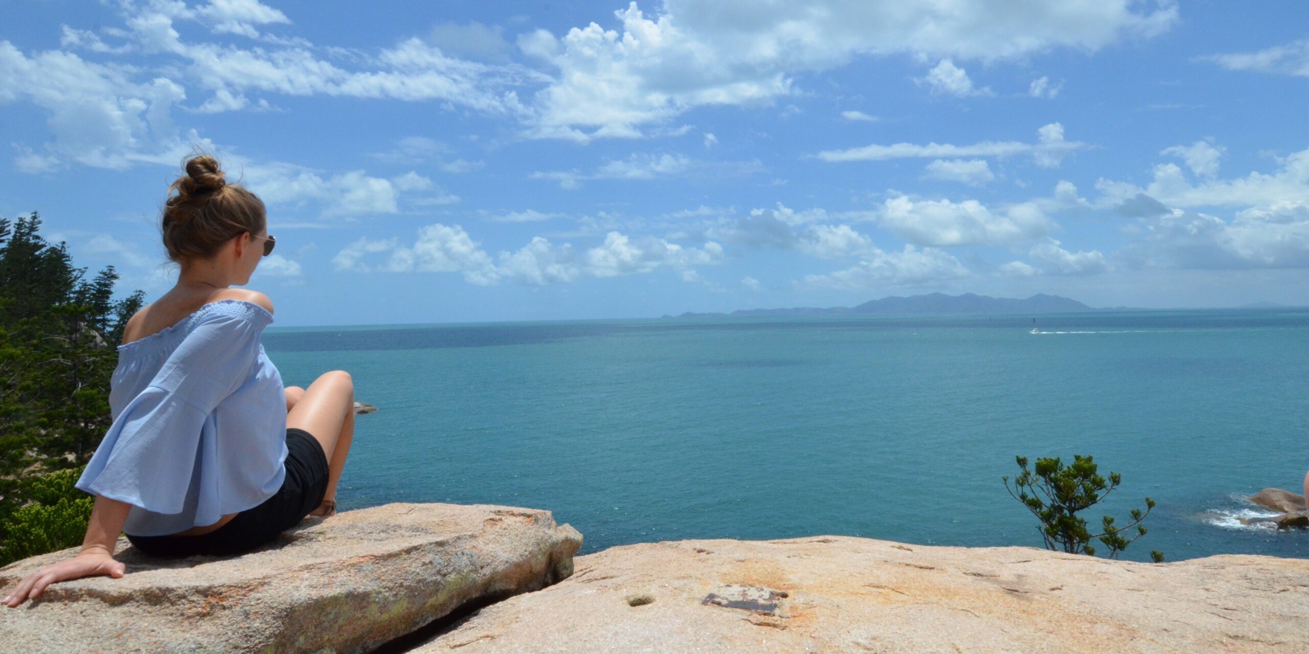 Po lewej siedząca tyłem na skale kobieta, w u dołu skała, w centralnej części ocean, u góry niebo z chmurami.